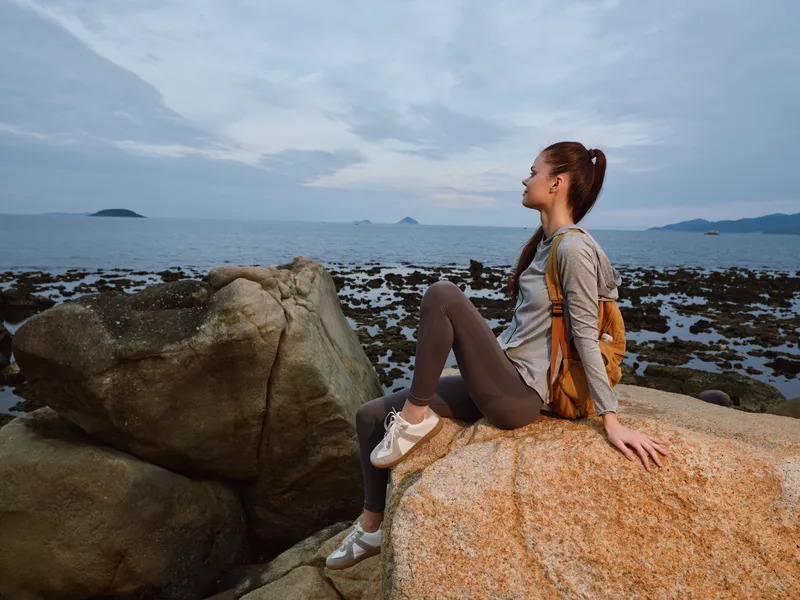 Young woman enjoying a serene moment on rocky beach at dusk, wearing casual sportswear and a backpack, surrounded by a tranquil sea landscape