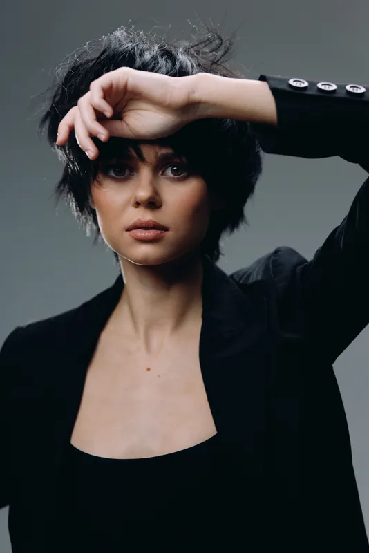 young woman with short black hair posing with hand near face wearing black blazer and dark top