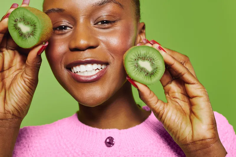 Smiling black woman with dark skin holds two kiwi halves near face on green background. She wears pink sweater and shows healthy teeth. Concept of fresh fruit, wellness, and natural beauty.