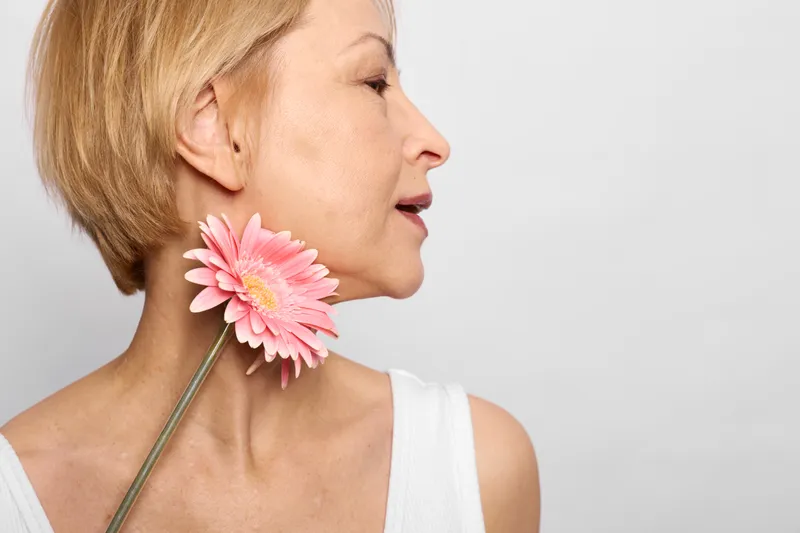 Woman portrait profile closeup, short blond hair, holding pink gerbera to her neck, calm smile, white sleeveless top, side view, soft light, minimal studio background