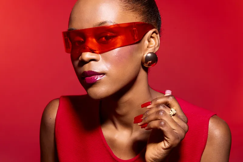 Stylish black woman with dark skin wearing red sleeveless top and futuristic red sunglasses poses against red background. Concept of fashion, beauty, confidence, and modern style.