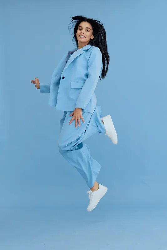 young woman jumping joyfully in blue suit and white sneakers against plain blue background, expressing happiness and energy in casual modern style