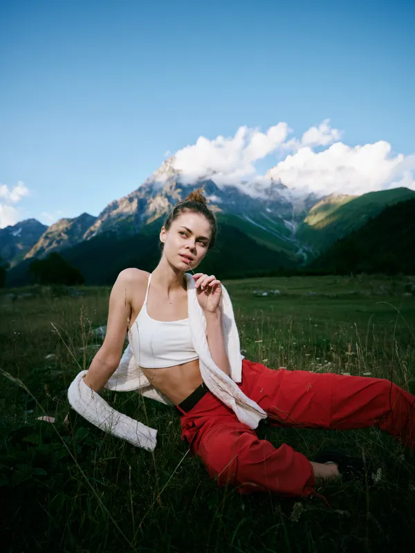 Active outdoor scene featuring a woman in a white sports top and red pants resting on grass with a towel, mountains and blue sky behind for a relaxed athletic moment