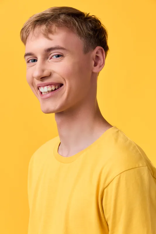 Smiling man in a yellow shirt stands in a bright studio, casual fashion, cheerful expression, bold color backdrop, warm lighting and approachable, friendly vibe.