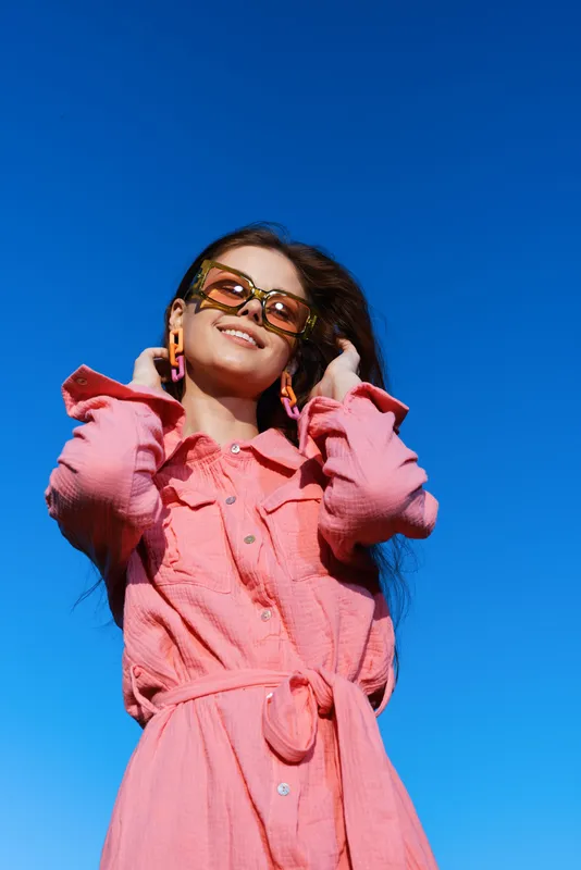 Young woman in a pink dress smiling against a clear blue sky, showcasing a vibrant summer fashion style Bright colors and confident emotions shine through