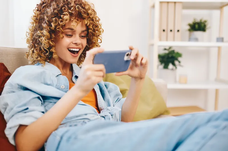 Уютный домашний отдых: женщина держит смартфон на диване, улыбается и наслаждается онлайн-играми