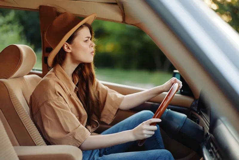 A young woman of European descent wearing a brown shirt and hat, driving a vintage car with a focused expression against a green outdoor background.