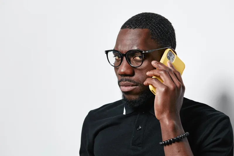 African American man with dark skin and glasses talking on yellow smartphone in casual black shirt against white background. Communication, technology and portrait with serious expression.