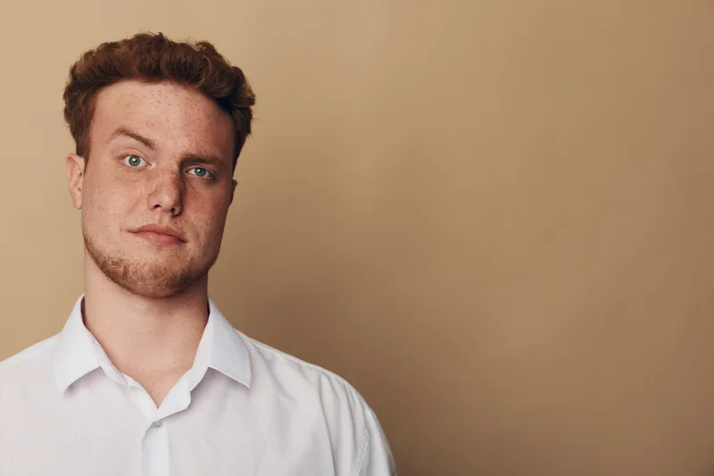 Young man with curly hair and beard wearing white shirt looks at camera with neutral expression on beige background. Concept of casual style, portrait, and natural appearance.