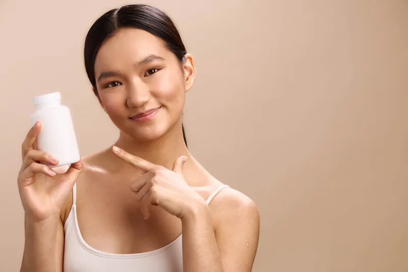 Asian woman holds a white bottle and smiles softly, presenting a cosmetic product in a clean studio. The calm pose and warm lighting convey trust, health, and personal care.