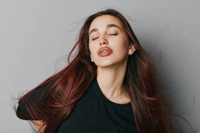 Young woman with long brown hair and closed eyes posing with relaxed expression in black shirt against neutral background. Beauty, calm, fashion, and natural makeup portrait in studio light.