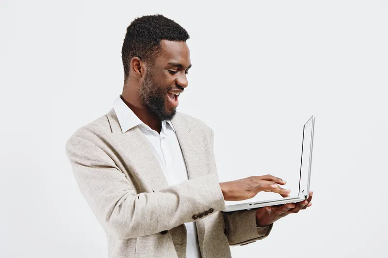 Smiling man wearing beige blazer and white shirt holding a laptop computer, looking at screen with a happy expression, isolated on white background, studio portrait, lifestyle concept.