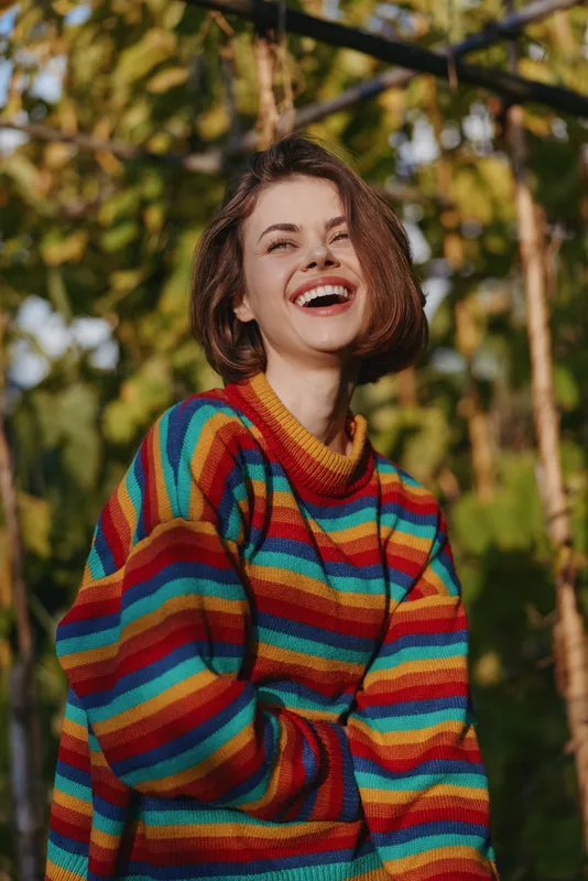 Woman smiling in a colorful striped sweater outdoors, young adult with short bob haircut laughing in a sunlit garden, casual knit fashion portrait conveying joyful, carefree lifestyle and seasonal