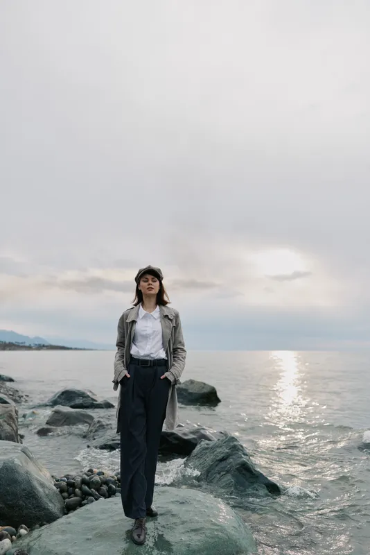 Young woman stands on rocky shore near calm sea under cloudy sky. She wears casual outfit and looks confidently ahead. Concept of nature, solitude, and peaceful moment outdoors.