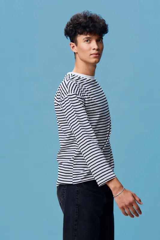 Young man with curly hair wearing a striped shirt and black jeans standing against blue background, looking sideways with a calm and composed expression.