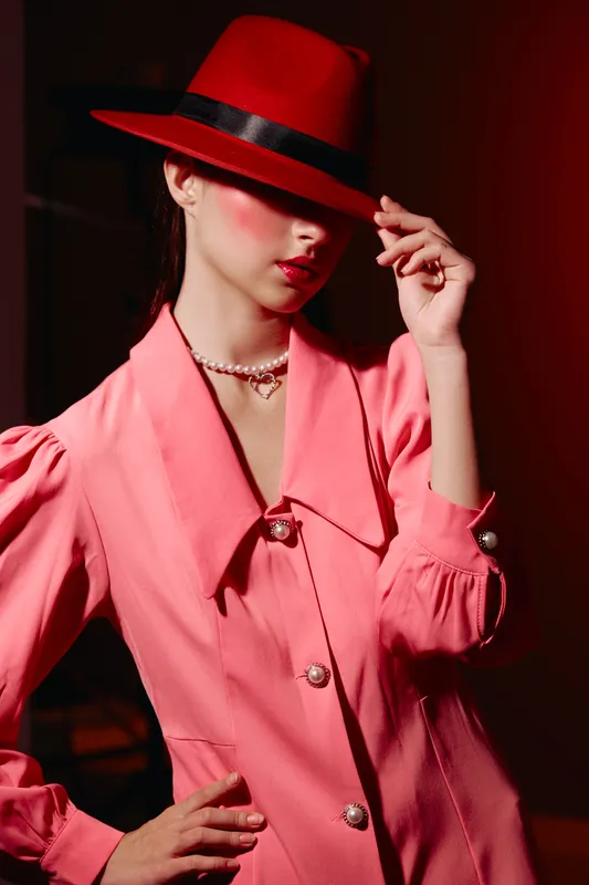 Fashion woman in pink dress and red hat poses dramatically adjusting hat brim. High-contrast lighting with red background emphasizes stylish beauty and creative fashion concept.