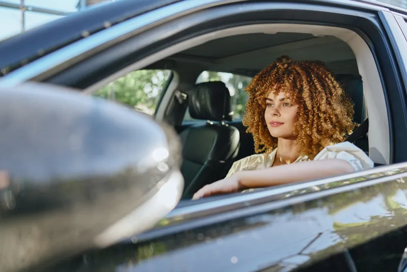 woman driving car with curly hair seated inside black vehicle on a sunny day with green blurred background