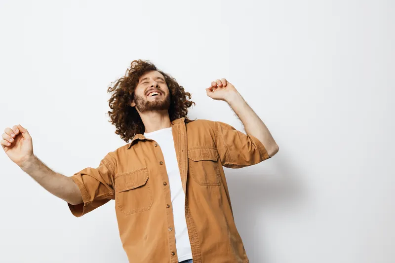 Happy young man dancing with curly hair in a casual shirt against a white background, expressing joy and positivity in a vibrant and uplifting scene