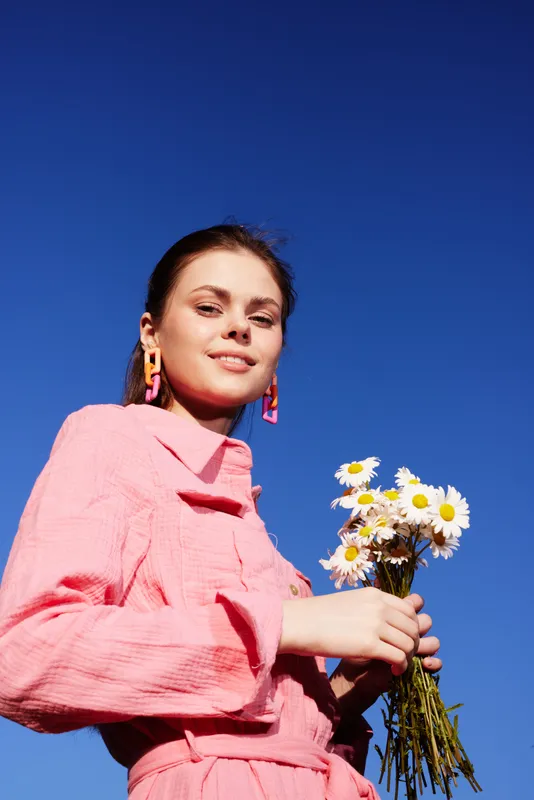 Young woman in a pink dress holding a bouquet of daisies against a bright blue sky, radiating joy and positivity Summer vibe with floral elements
