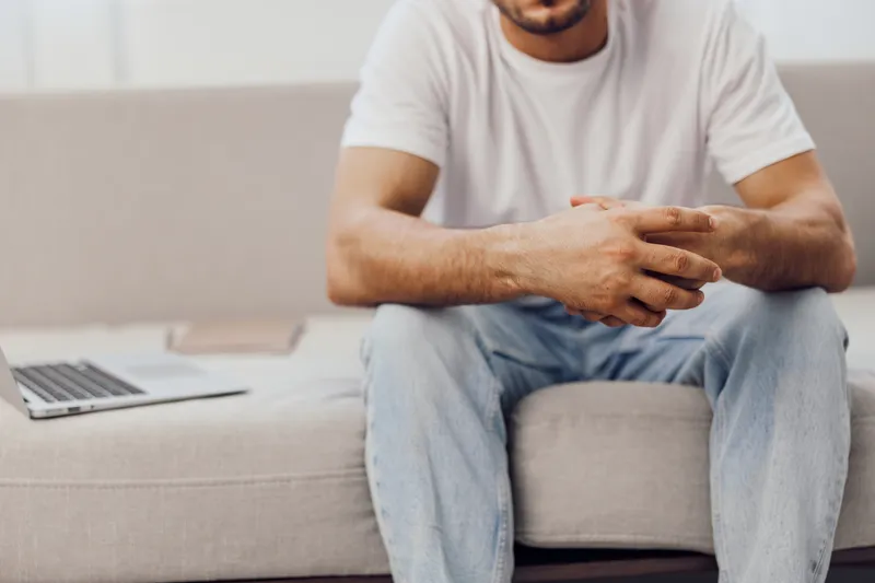 Casual man sitting on a sofa, appearing relaxed and thoughtful while engaging with a laptop. Ideal for themes of productivity and remote work in modern lifestyle.