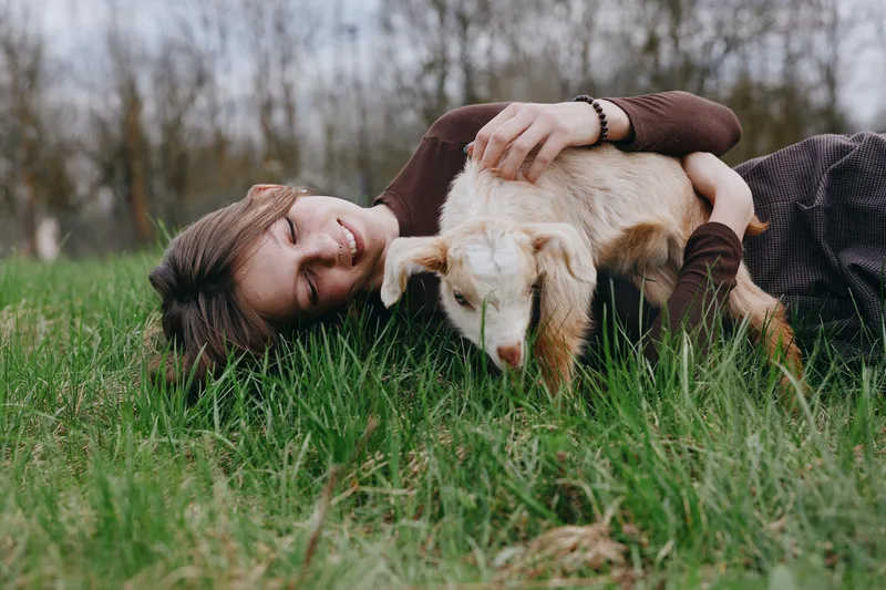 Warmhearted woman relaxing with a goat kid on a grassy field, outdoors lifestyle moment, gentle interaction and affection, natural light, candid mood.