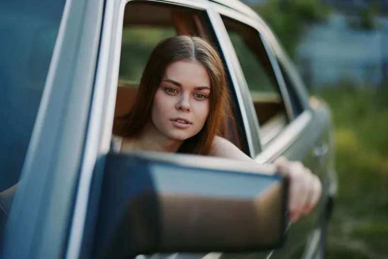 Young woman with long hair gazing out of a car window, expressing a sense of freedom and adventure in a natural green background, perfect for travel themes.