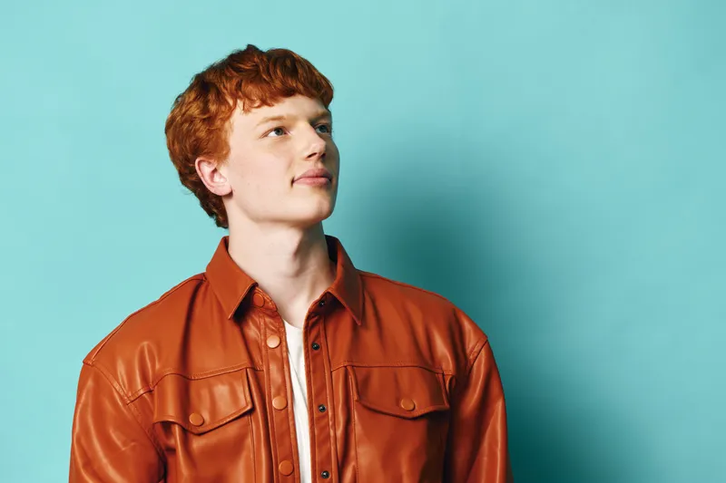 Young man with red hair wearing orange leather jacket standing against blue background looking away thoughtfully with confident expression.
