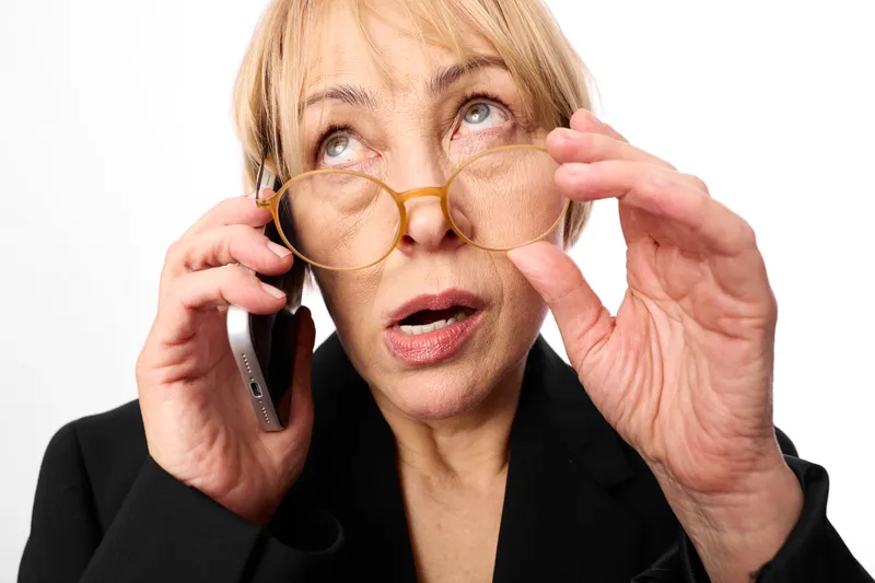 Surprised elderly woman in studio setting adjusts glasses while holding a phone to her ear, looking forward, focused on a call, communication