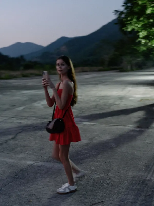 young woman in red dress holding smartphone standing on empty road at dusk, blurred background with mountains and trees, casual evening outdoor scene