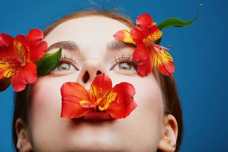 Beautiful young woman with green eyes surrounded by vibrant red flowers on a vivid blue background, expressing serenity and nature s elegance