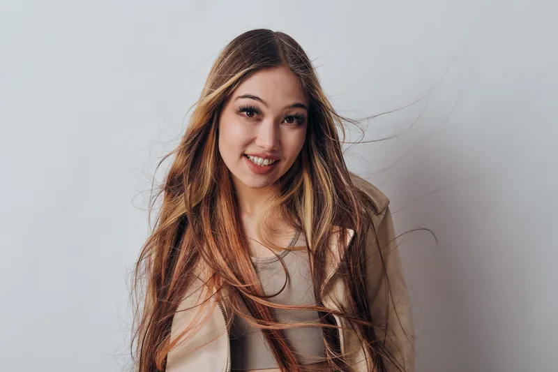 Young woman with long hair and natural makeup smiling against neutral background. Female model wearing casual beige jacket and top in studio portrait with joyful expression.