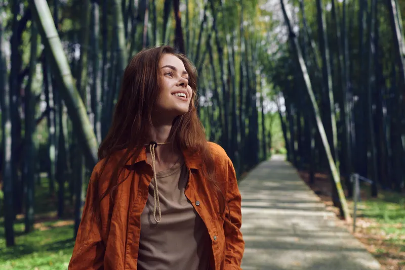 Woman portrait smiling while walking on a bamboo path outdoors in nature, casual redhead in jacket enjoying a serene stroll, candid lifestyle photo of walking and relaxation.