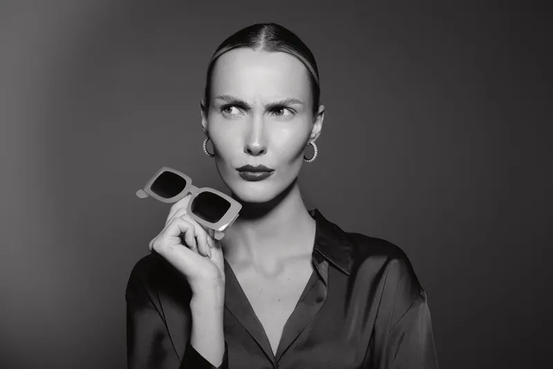 Young white woman with light skin holding sunglasses looking confused or skeptical in studio portrait. Concept of doubt, fashion, and expression in black and white photography.
