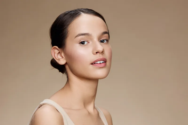 Young woman with natural makeup and a neutral expression, standing against a soft beige background, embodying beauty, confidence, and minimalism in a serene setting