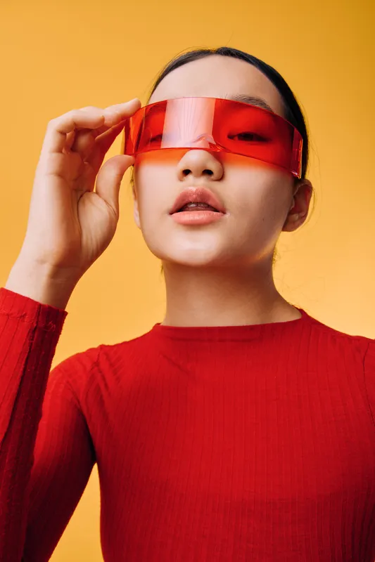 Woman wearing futuristic red visor glasses and red top, posing on yellow background with hand adjusting eyewear for fashion portrait and modern style concept.