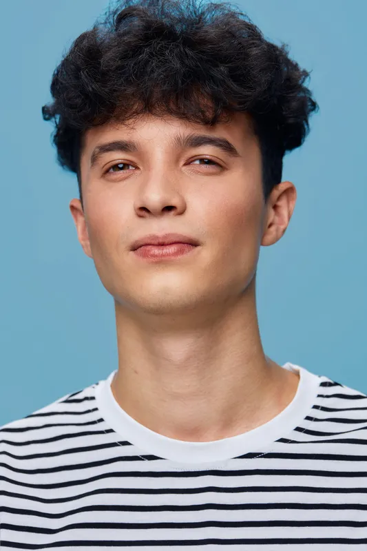 young man with curly hair wears casual striped shirt looking confident against simple blue background in studio portrait with soft lighting