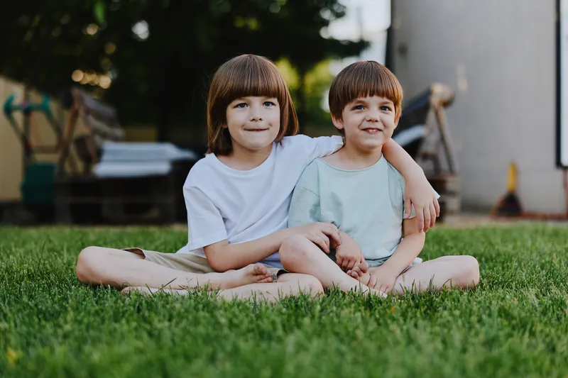 Two smiling boys enjoy a sunny day outdoors, sitting together on grass, dressed casually. Their cheerful expressions reflect happiness and friendship.