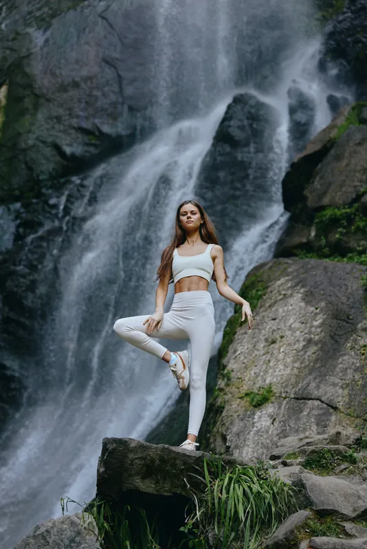 young woman practicing yoga in tree pose by a waterfall, wearing white activewear, peaceful outdoor meditation, natural green environment, serene balance