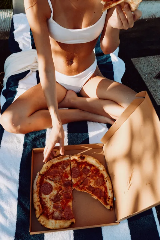 A young woman in a white bikini sits on a striped blanket outdoors, enjoying a slice of pizza from a box on a sunny day, radiating casual carefree mood and summer vibes