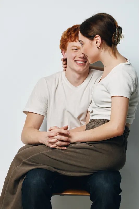 Young couple sitting close together, woman on man's lap with affectionate smile, casual clothing and relaxed posture at light neutral background.