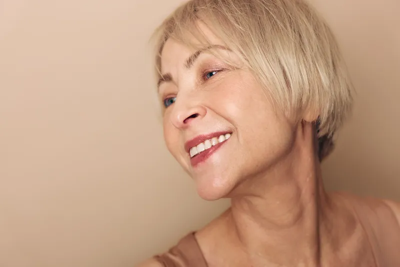 Smiling mature woman with short blonde hair looks away on beige background. Concept of happiness, aging, natural beauty, confidence, and positive emotions in lifestyle portrait.