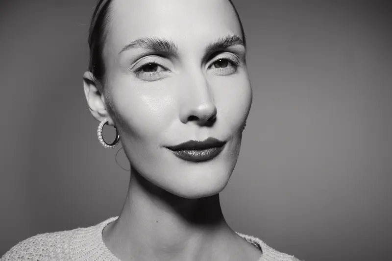 Close-up portrait of a white woman with fair skin and slicked-back hair wearing hoop earrings. Studio lighting highlights natural beauty and confident expression.