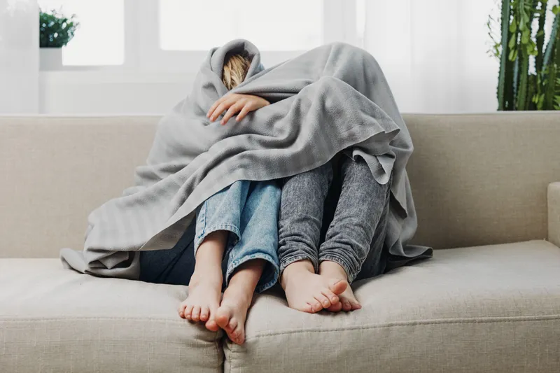 Two people sitting on a sofa wrapped together in a grey blanket, showing cozy and intimate moment at home with casual clothes and bare feet visible.