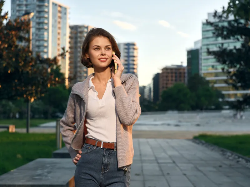 A stylish woman talks on a mobile phone in a modern urban park. She smiles warmly, wearing a cardigan and jeans, projecting confidence and a relaxed professional vibe.