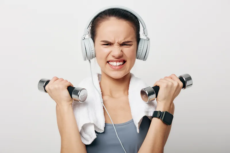 Woman fitness enthusiast lifts dumbbells with headphones and towel, showcasing focus and determination during a gym workout, a strong sport and exercise moment in bright studio.