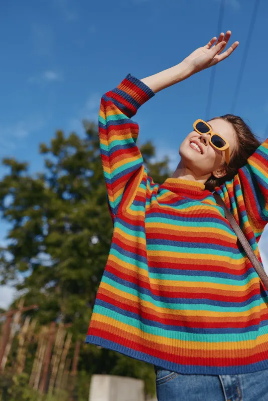 woman rainbow sweater sunglasses smiling outdoor portrait casual outfit joy, young woman in oversized striped knit stretching arms up under blue sky with pine trees, happy lifestyle fashion travel