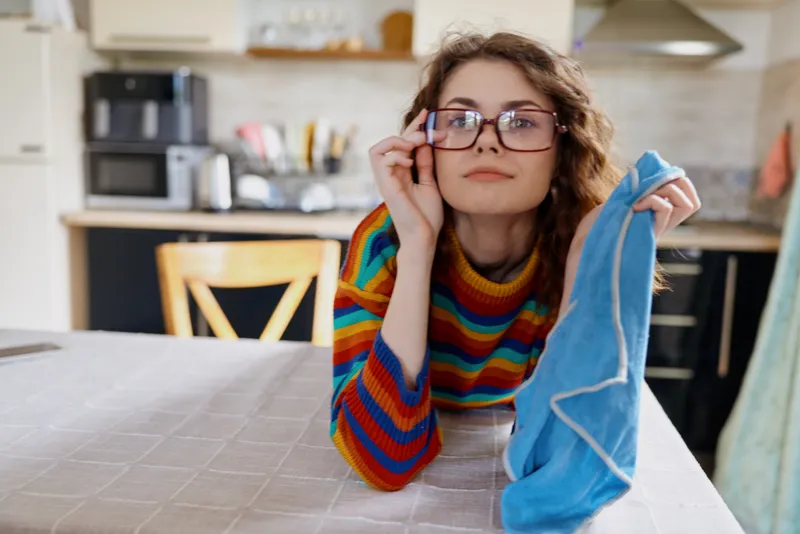 Young woman with curly hair and glasses sits at a cozy kitchen table, holding a blue sock and peering over the rim with a curious, playful smile.