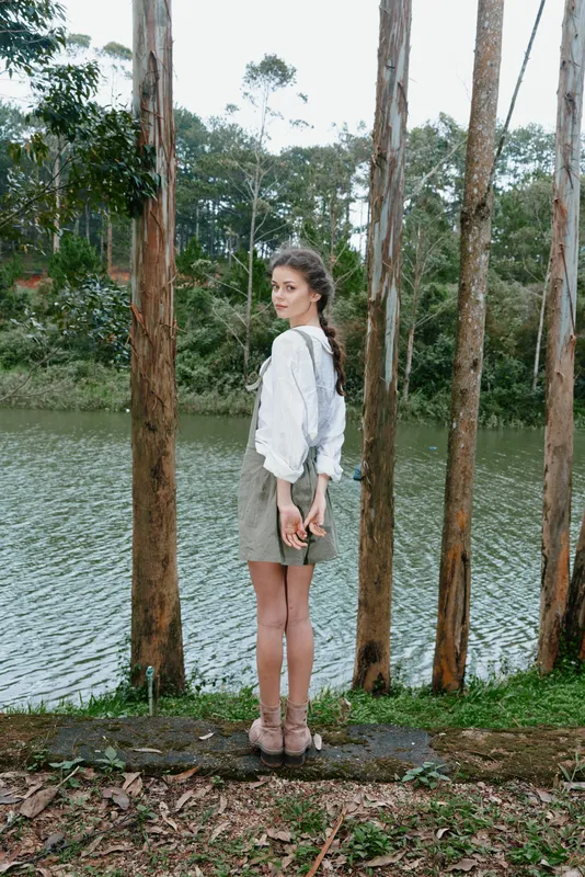 Young woman in casual dress by the lake, expressing tranquility among nature, wearing a light sweater and shorts, with a serene background of trees and water