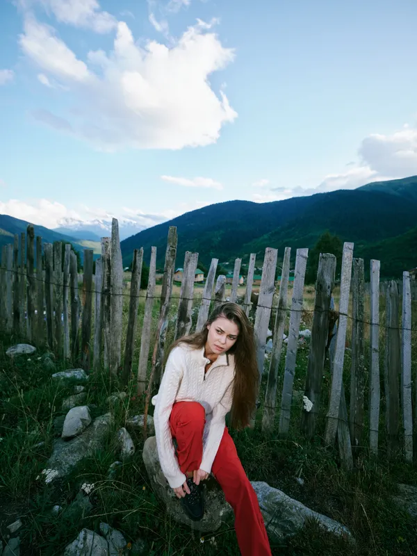 Woman sits by a weathered wooden fence in a mountainous landscape, wearing a light sweater and red pants, exploring nature and countryside ambience under a blue sky with scattered clouds.