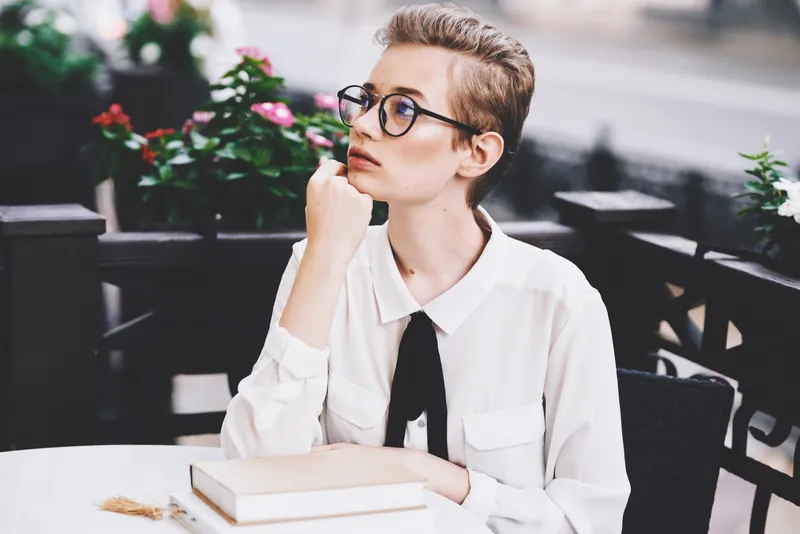 Молодая женщина в рубашке и очках с книгой за столиком в кафе
